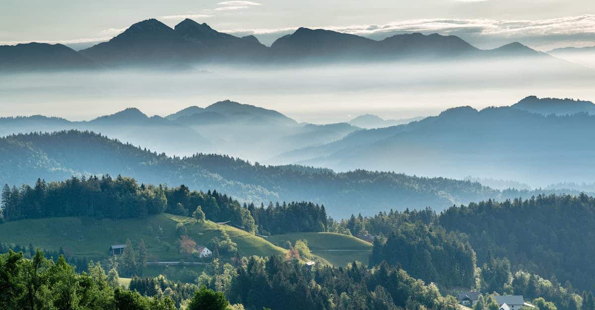 A scenic view of rolling hills covered in trees with scattered houses, leading to distant misty mountains under a partially cloudy sky.